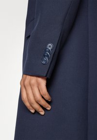 Marinblå kavajärm med fyra mörka knappar med logotyp, texturerad tyg och en skräddarsydd design. Handen vilar vid manschetten.