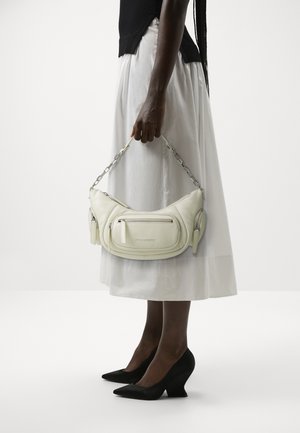 Personne portant une jupe blanche et un haut noir tenant un sac beige clair avec plusieurs poches zippées et une anse chaîne.