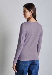 Maglietta a maniche lunghe in cotone lavanda con scollo rotondo, abbinata a jeans in denim blu. Tessuto morbido, taglio aderente, dettagli di cucitura visibili.