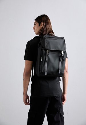 Crni najlon ruksak s gornjom preklopom, podesivim trakama i zippered prednjim džepom. Sadrži logo na preklopu i pravokutni oblik.