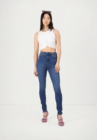 Bílý krátký top s uzlovým detailem, modré džíny s vysokým pasem a fialové sandály na podpatku se šňůrkami. Hladká látka a přiléhavý střih.