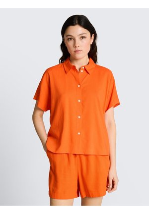Femme aux cheveux foncés portant une chemise boutonnée orange vif à manches courtes et un short assorti sur un fond clair uni.