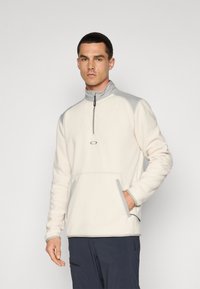 Lys kremfarget fleece-genser med halv glidelås i kragen, grå detaljer på skuldrene og lommekantene, samt en frontlomme i kenguru-stil.