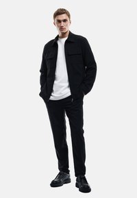Schwarze Jacke mit zwei Taschen auf der Vorderseite, getragen über einem weißen T-Shirt. Schwarze Hose und Schuhe runden das Outfit ab. Einfaches, modernes Design.