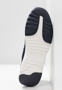 Granatowe sneakersy z teksturowaną siateczką na górze, z kremową gumową podeszwą o wzorzystym gripie i wypukłych akcentach, ustawione pod kątem na białej powierzchni.