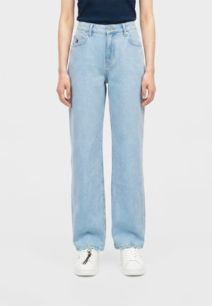 Personne portant un jean droit bleu clair et des baskets blanches, debout devant un fond uni et clair.