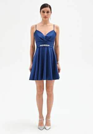 Femme debout portant une robe en satin bleue sans manches avec bretelles fines, détail orné à la taille, et des escarpins argentés.