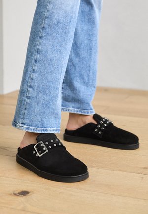 Fekete velúr slip-on cipők lapos talppal, széles pánttal, ezüst színű fémrészekkel és díszes szegecsekkel. Világoskék farmerral viselve.
