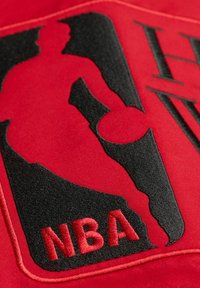 Tela roja con un bordado del logo negro de la NBA, que representa a un jugador y las letras "NBA" en rojo. El diseño tiene una textura suave.