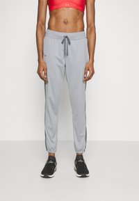 Personne athlétique portant un crop top rouge, un pantalon de jogging gris clair avec cordon de serrage et des baskets noires, debout devant un fond blanc uni.