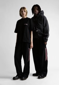 Camiseta negra de gran tamaño, de manga corta, con un logo bordado en rojo. Sudadera negra con cremallera en el frente, pantalones holgados negros con detalles del logo.