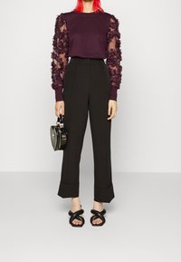 Donna con capelli rossi vivaci che indossa un top viola scuro con maniche trasparenti e volantes, pantaloni neri a vita alta, sandali neri con cinturini intrecciati, mentre tiene una piccola borsa nera.