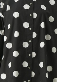 Chemise noire avec de grands pois blancs, devant boutonné avec de petits boutons verts, et une texture de tissu douce et lisse.