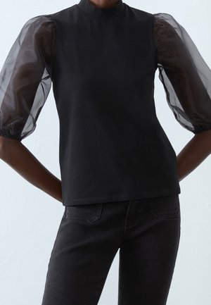 Femme portant un haut noir avec des manches bouffantes transparentes et un col montant, associé à un jean noir en denim, debout les mains sur les hanches.