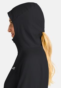 Zwarte waterdichte jas met een aansluitende capuchon, met een gladde textuur en contouren. Logo zichtbaar nabij de zoom. Haar dat uit de capuchon valt.