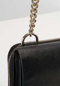 Sort struktureret læderhåndtaske med en gylden kædetræk og lynlåsdetailler. Har en rund form og en metalring til fastgørelse.