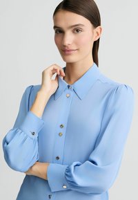 Svetle modrá blůza z průhledné látky, s klasickým límečkem, přední částí na knoflíky se zlatými knoflíky a nadýchanými dlouhými rukávy.
