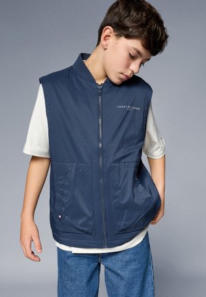 Námornícky modrá vesta s vysokým golierom, predným zipsom a dvoma veľkými vreckami. Na hrudi sa nachádza logo "Tommy Hilfiger". Ľahký materiál.