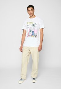 Hvid bomulds t-shirt med et farverigt grafisk design af planter og arkitektur, parret med beige afslappede bukser og sneakers.