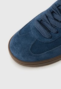 Μπλε σουέντ αθλητικό παπούτσι με υφή, με ταιριαστά επίπεδα κορδόνια και καφέ λαστιχένια σόλα, που παρουσιάζει καμπυλωτά σχέδια.