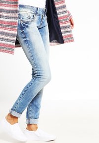 Ljust blå denimjeans med slim fit, lätt slitna detaljer och uppvikta fållar, bärs med vita slip-on skor och en mönstrad kappa.