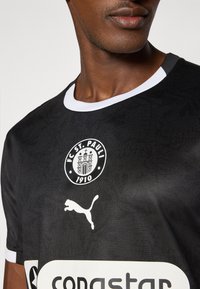 Maillot de football noir avec un col rond blanc, logo du FC St. Pauli, logo Puma en blanc, et le nom d'un sponsor imprimé sur la poitrine.