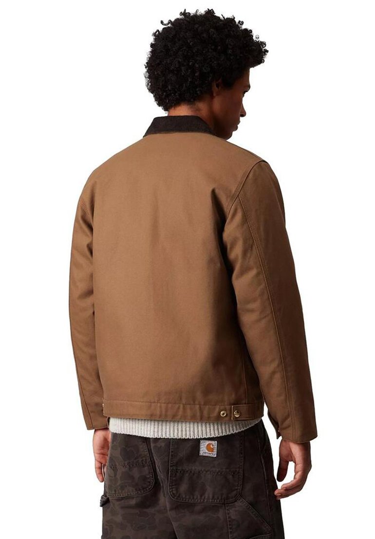 Man with curly hair wearing a brown jacket over a white knit sweater and dark patterned cargo pants, facing away against a white background.