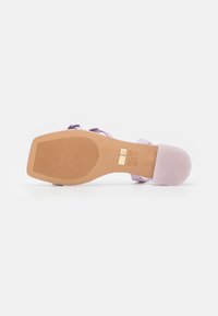 Sandale violette à lanières avec une semelle lisse et texturée. Dispose d'un talon carré et de rainures verticales pour une meilleure adhérence. Accent avec logo doré sur la semelle.