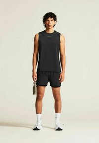 Junger Mann in schwarzem ärmellosem Shirt und Shorts, der eine metallische Wasserflasche hält, weiße Socken und Sportschuhe trägt und vor einem schlichten Hintergrund steht.