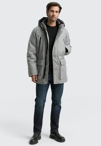 Grå, polstret jakke med aftagelig sort hætte, lynlås foran og flere lommer. Båret over en sort sweater og blå jeans.
