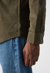 Olivengrønn kordfløyelskjorte med rett snitt, knappede mansjetter og en sidelapp; kombinert med lyseblå denimjeans, som viser hånddetaljer.