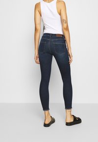 Jeans skinny azul oscuro con bolsillos traseros, ligera deshilachadura en el dobladillo, combinados con chanclas negras. Tejido de mezcla de algodón con un acabado suave.