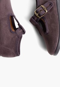 Zapatos de ante púrpura oscuro con una textura suave, lados abiertos y un acento de hebilla dorada. Diseñados con una forma redondeada y suela minimalista.