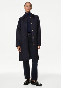 Trench coat navy con cuello tipo solapa, cierre de botones y mangas largas. Estilizado con jeans oscuros y mocasines negros, sobre un fondo blanco.