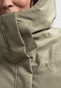 Beige wasserdichte Jacke mit hohem Kragen, verziert mit Tropfen. Verfügt über ein dezentes Logo und einen strukturierten Stoff mit glatter Oberfläche.