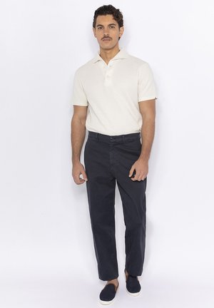 Polo crème, à manches courtes, avec col, associé à un pantalon marine, à coupe droite et légèrement brillant. Chaussures bleu foncé.