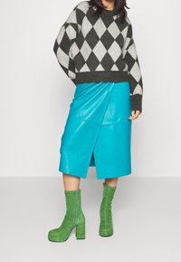 Maglione argyle bianco e nero lavorato a maglia, gonna avvolgente in pelle blu lucida e stivali alla caviglia in suede verde con tacco a blocco.