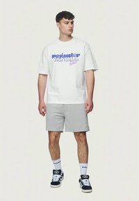 Bílá tričko s fialovým textem "Predator Vanilla", v kombinaci se šedými kraťasy. Model má na sobě černé tenisky a bílé ponožky s logem.