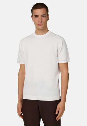Jeune homme aux cheveux courts portant un t-shirt blanc uni à manches courtes et un pantalon foncé, sur un fond uni.