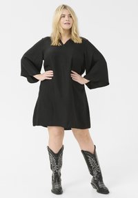 Robe noire avec un col en V et des manches larges, fabriquée en tissu texturé. Associée à de grandes bottes de cowboy noires avec des accents argentés.