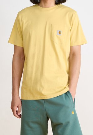 Person iført en gul Carhartt-lomme T-shirt og turkise Carhartt-bukser med den ene hånd i lommen mod en hvid baggrund.