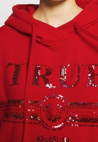 Červená mikina s kapucí se třpytivým logem "TRUE RELIGION", obsahující kulatý emblém a vodorovné pruhy, která představuje hladkou, jemnou texturu.