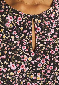 Gros plan d'un tissu noir à motif floral avec de petites fleurs roses, jaunes et blanches, présentant une encolure en trou de serrure avec un seul bouton noir.