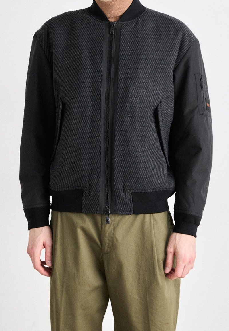 Blouson bomber noir avec un corps texturé, des manches lisses, une fermeture éclair sur le devant et des poignets côtelés. Comprend une poche zippée sur la manche.