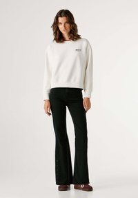 Biely mikina s rebrovanými manžetami, s malým vyšitým logom. Kombinovaná s čiernymi rozšírenými džínami, detailom s gombíkom na leme a maroon teniskami.