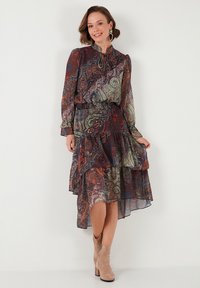Langärmeliges, paisley-mustertes Kleid in Dunkelblau, Rot und Grün, mit einem rüschend geschnittenen Saum und Halstie. Kombiniert mit beigen Knöchelstiefeln.