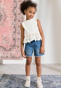 Weißes ärmelloses Oberteil mit Spitzenabschluss, kombiniert mit Jeansshorts und weißen High-Top-Sneakern. Im Hintergrund ist ein rosa gemusterter Teppich zu sehen.