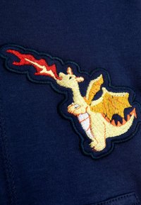 Parche bordado de dragón sobre tela de color azul oscuro, con un dragón amarillo escupiendo fuego, y con un detallado bordado en naranja y negro.