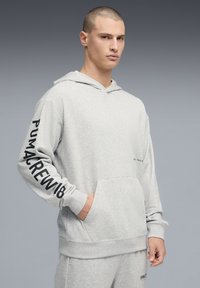 Sweat à capuche gris en tissu doux, avec une poche avant, une capuche ajustable et un texte noir sur la manche. Design simple avec une coupe décontractée.