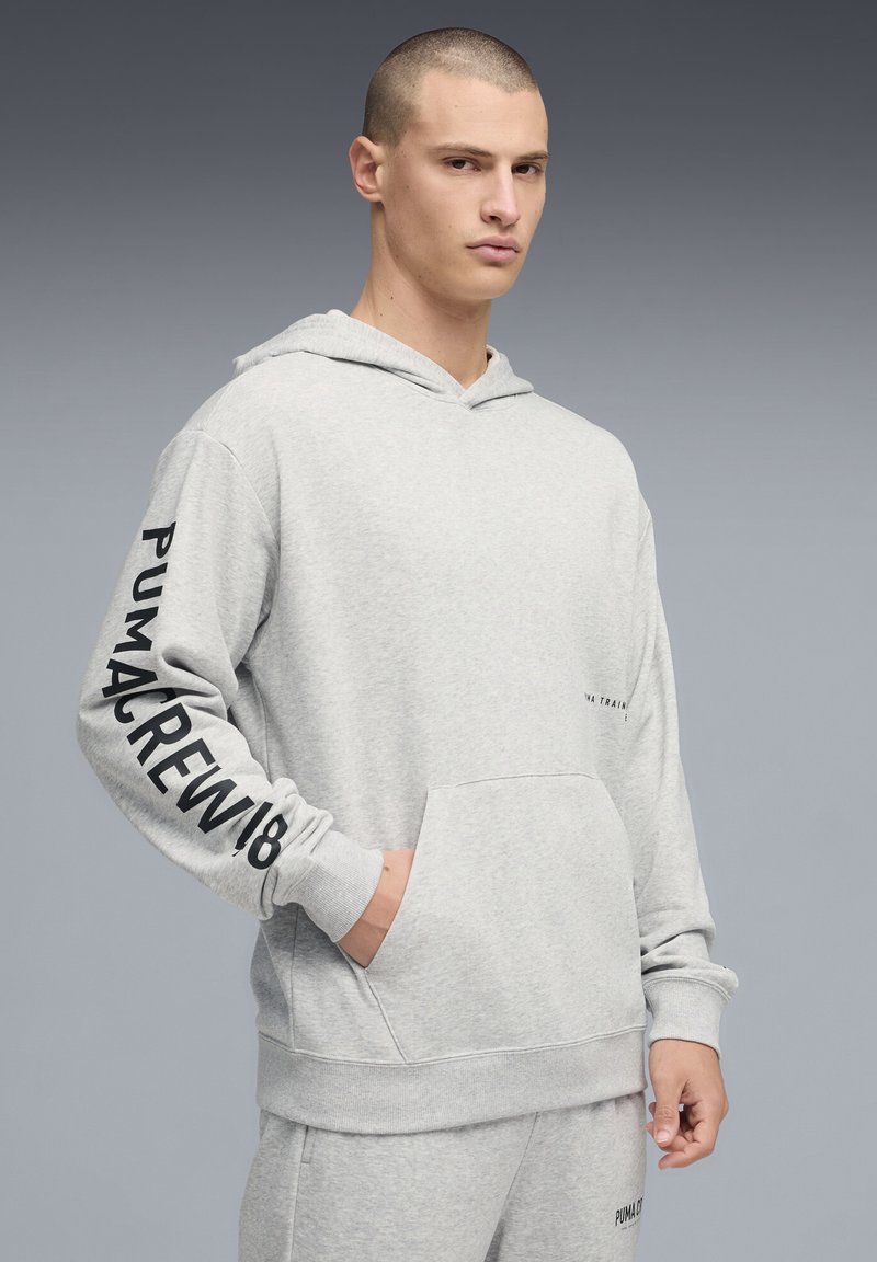 Sweat à capuche gris en tissu doux, avec une poche avant, une capuche ajustable et un texte noir sur la manche. Design simple avec une coupe décontractée.